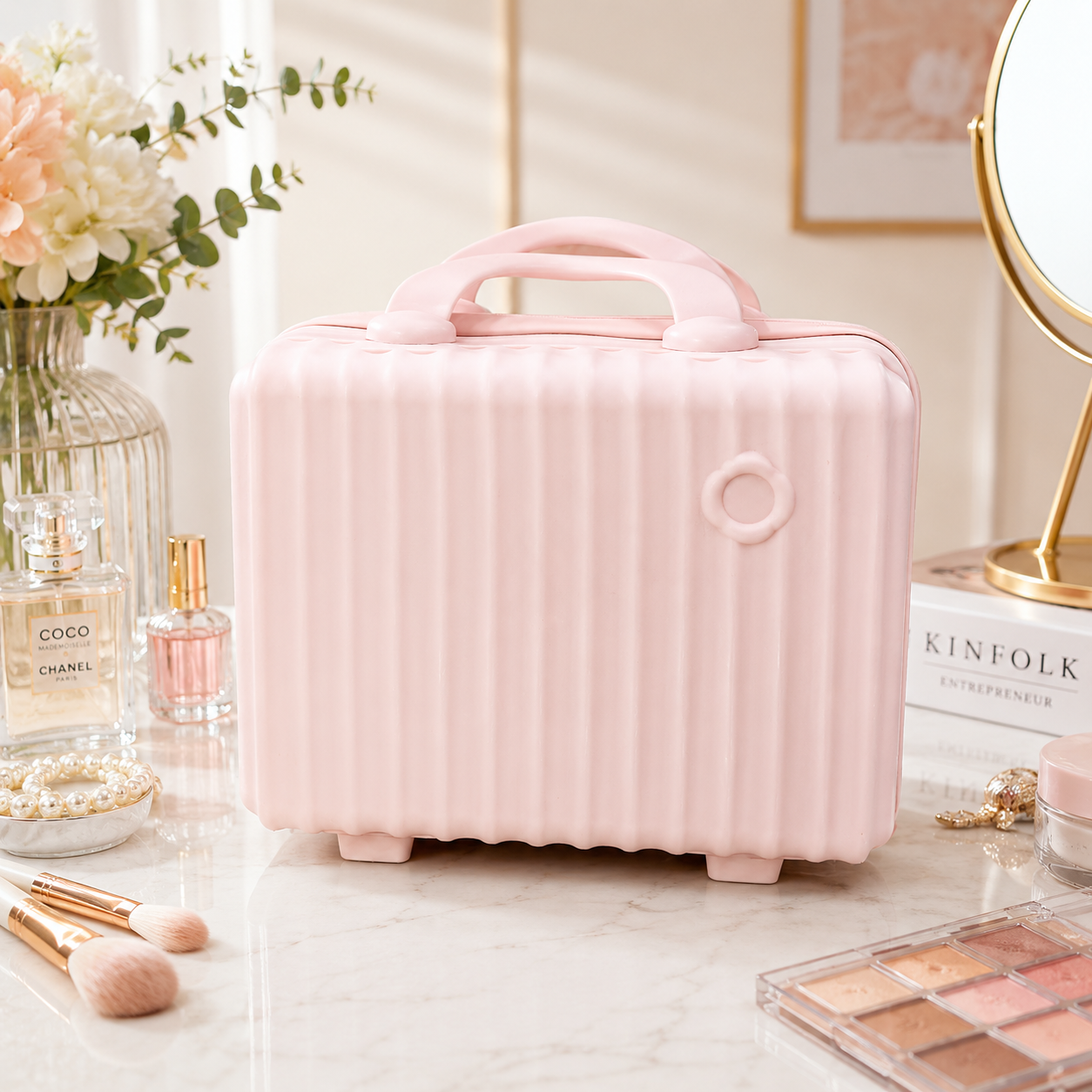 A light pink cosmetic case designed like a small suitcase with a top handle, standing on a marble tabletop surrounded by beauty props including perfume bottles, makeup brushes, a mirror, and an eyeshadow palette.