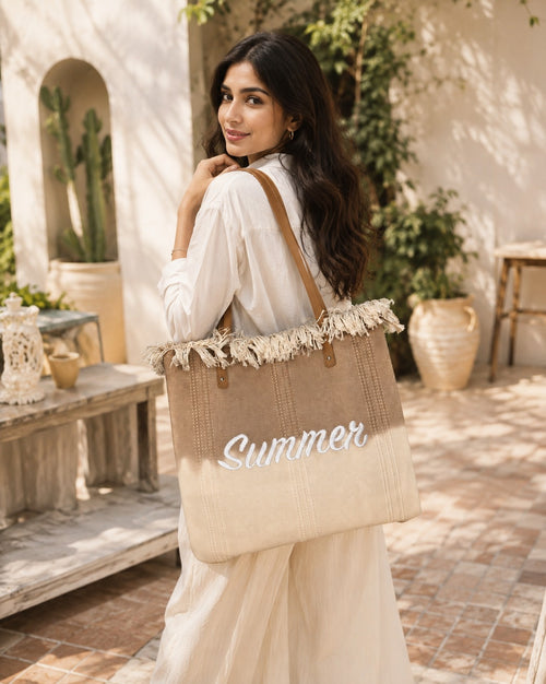 A woman in a white dress carries a large beige woven tote bag with fringe trim and the word 'Summer' printed on the front, set in a sunlit outdoor courtyard.Summer Jute Tote Bag for Women – Large Beach & Vacation Handbag